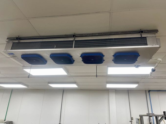 Ceiling-mounted industrial cooler with four blue air vents in a white-walled room.