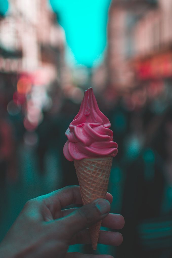 Hand holding a pink ice cream cone in a busy street, blurred background.