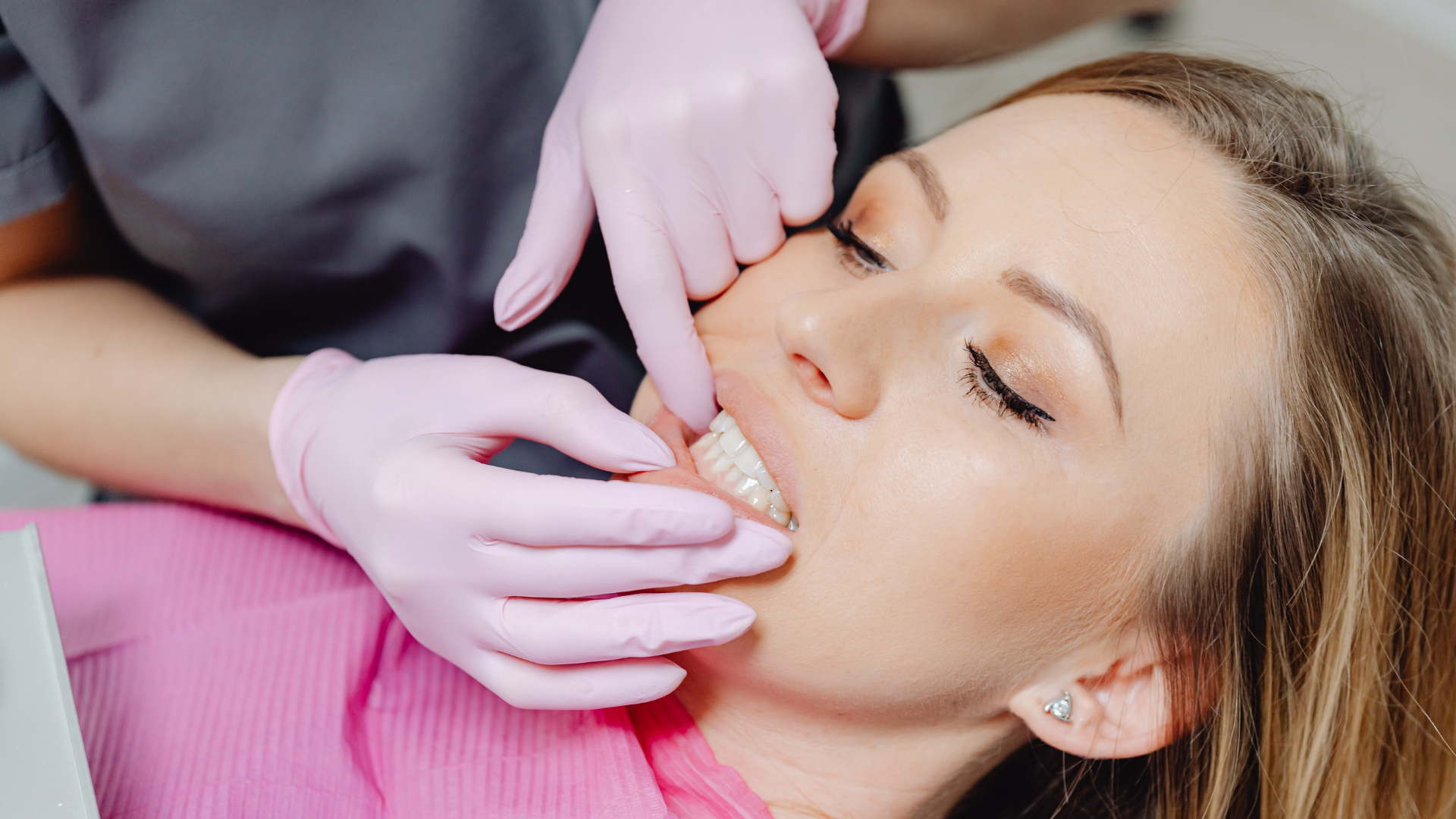 Dentist's gloved hands near patient's mouth, examining clear dental aligner. Patient reclined, eyes closed.
