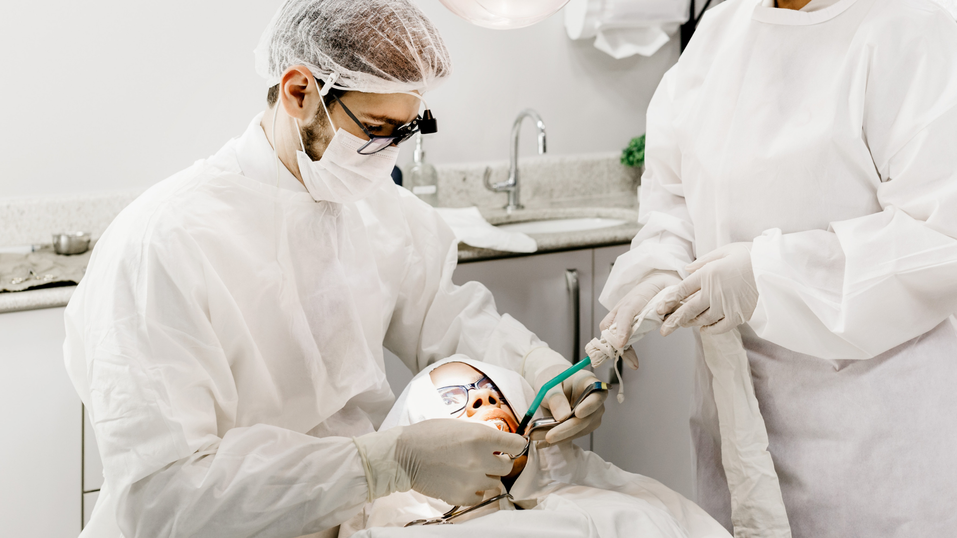 Dentist in protective gear examining a patient's mouth with an assistant in a dental office.