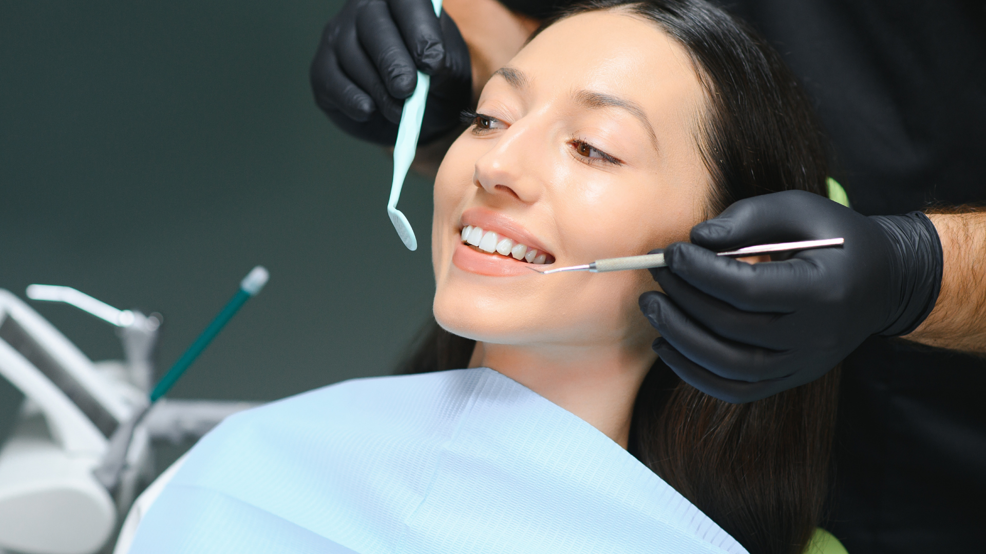 Woman at the dentist, smiling while teeth are being examined with dental tools, in a medical setting.