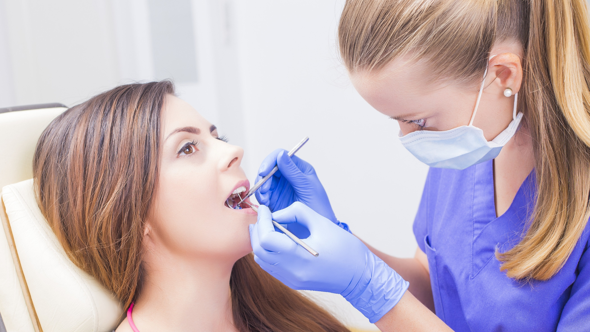 A dental professional in blue scrubs and a face mask examines a patient's teeth with a mirror and explorer.