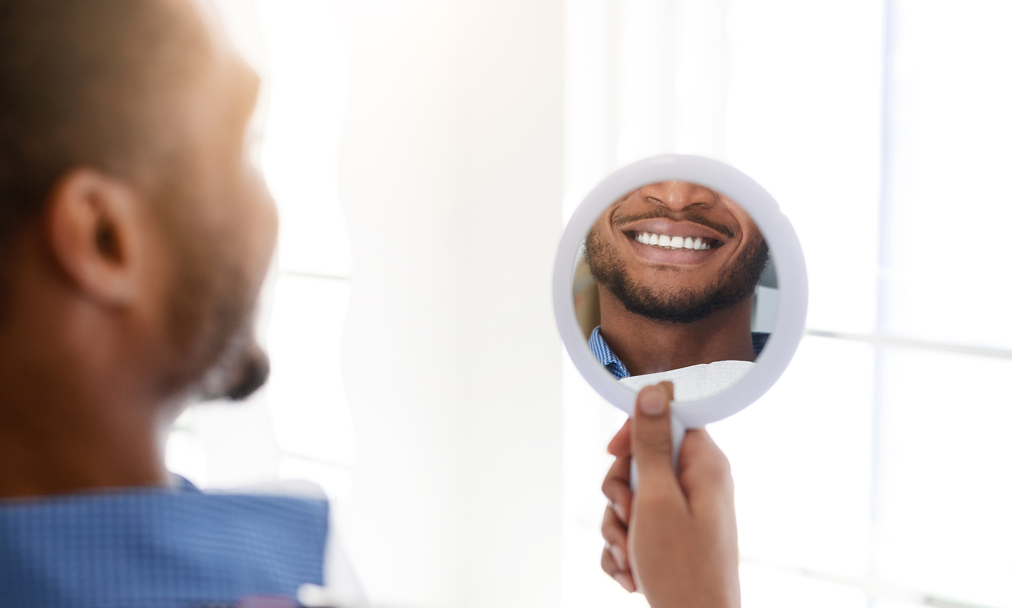 A man is looking at his smile in a hand mirror.