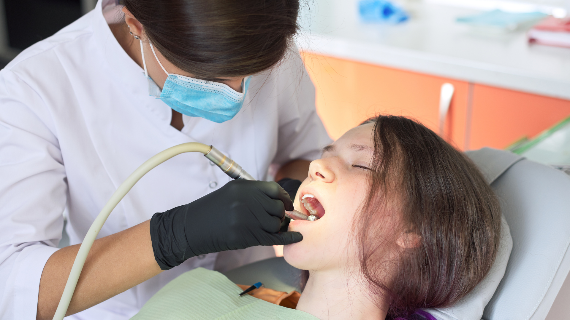Dentist examining a patient’s mouth with tools, both wearing masks and gloves, in a clinic chair.