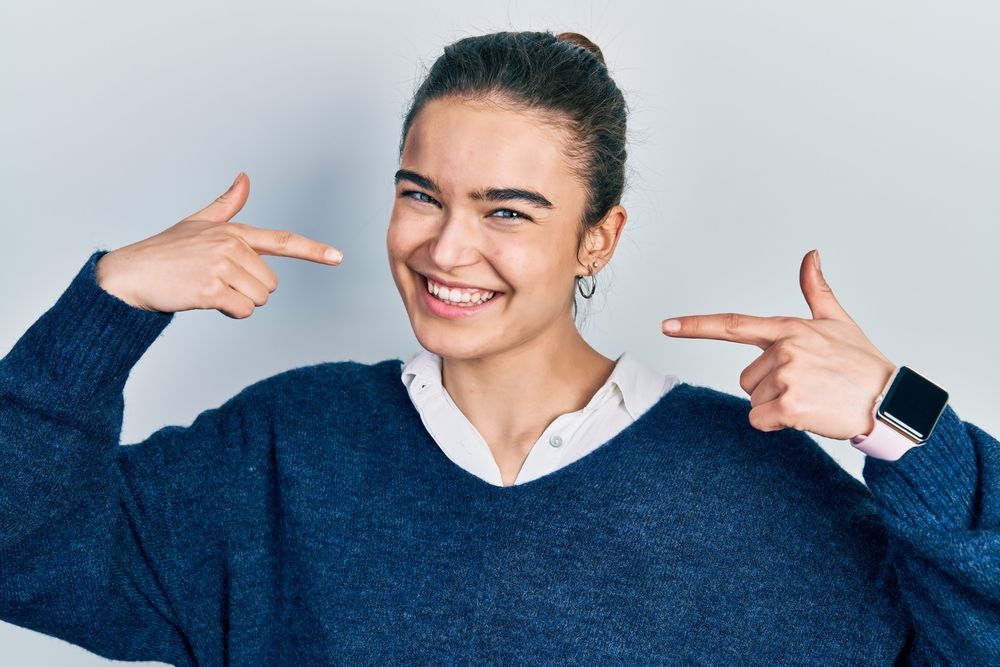 A young woman in a blue sweater is smiling and pointing at herself with both hands.