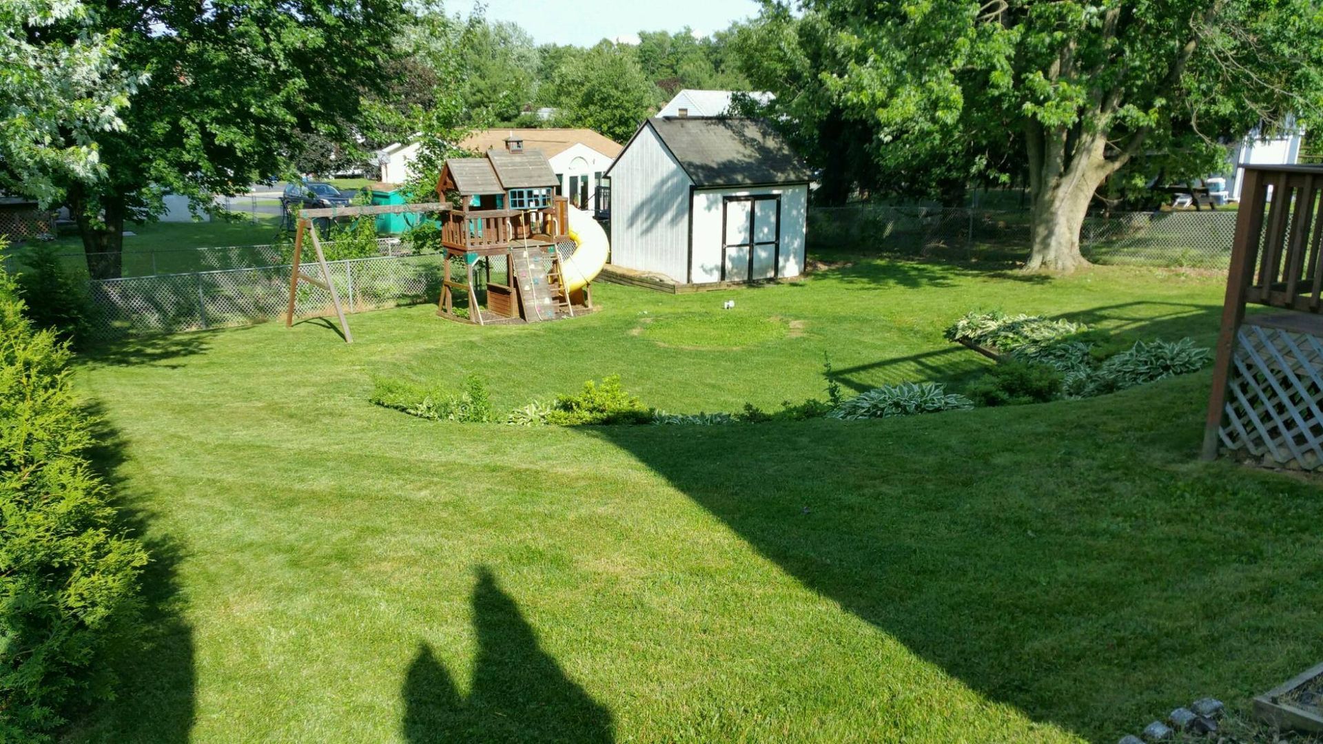A grassy backyard featuring a small white shed, a children's play set, a wooden deck, and mature trees under a sunny sky.