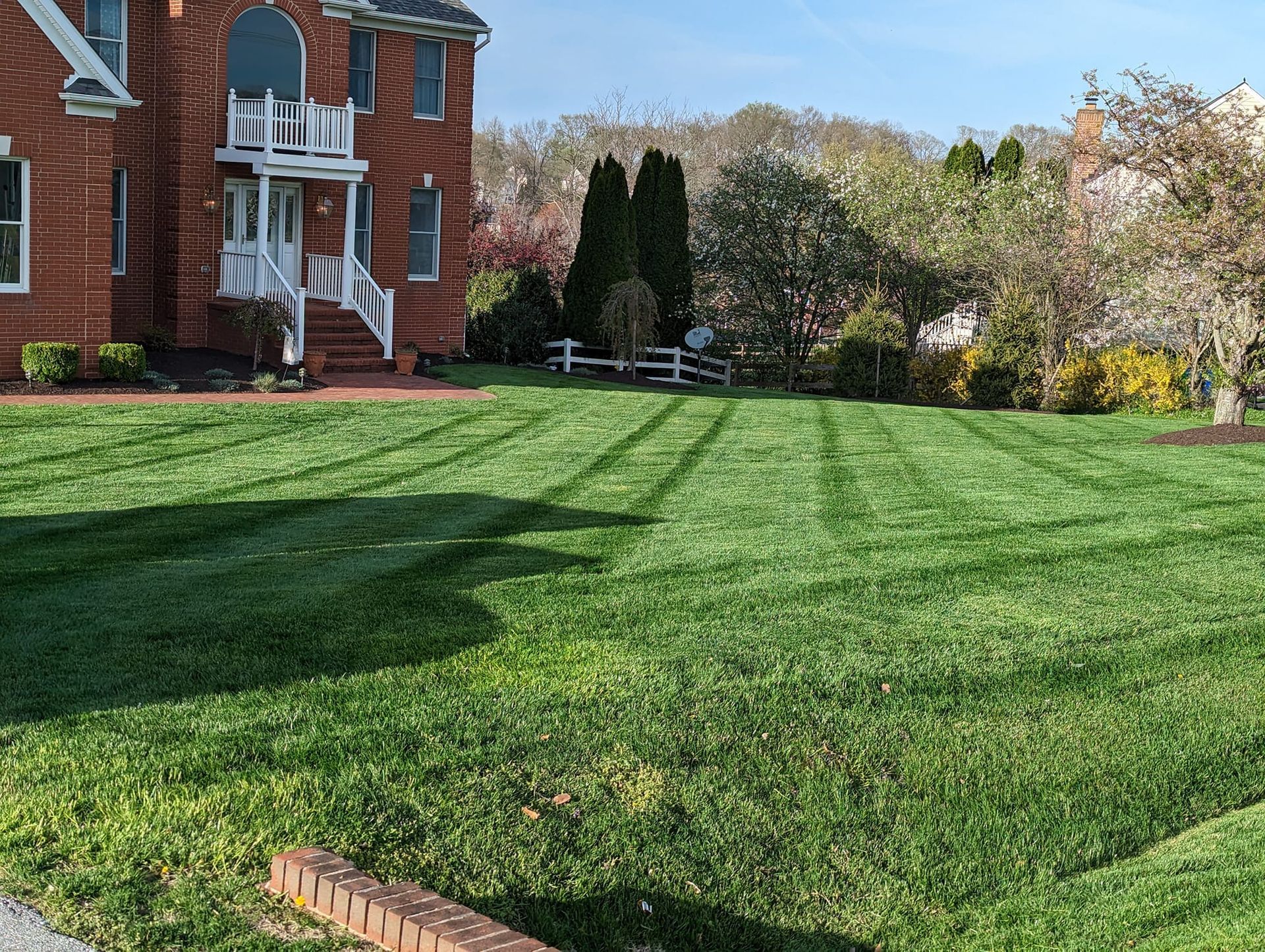 A lush green lawn with clear mowing stripes in front of a brick house under a blue sky.