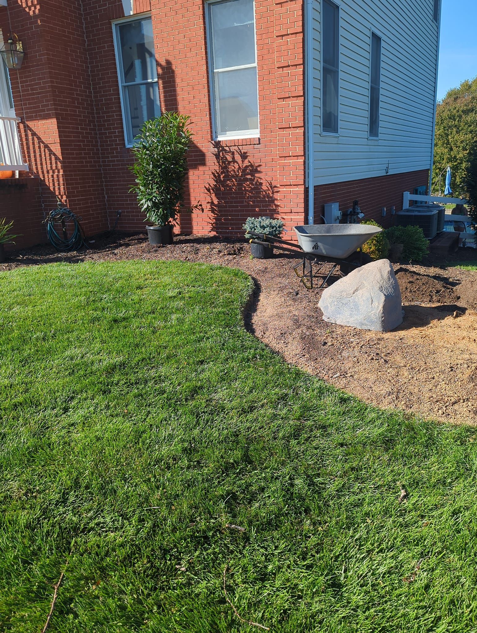 A manicured lawn curves around a flower bed with mulch, a large boulder, and potted plants near a red brick building.