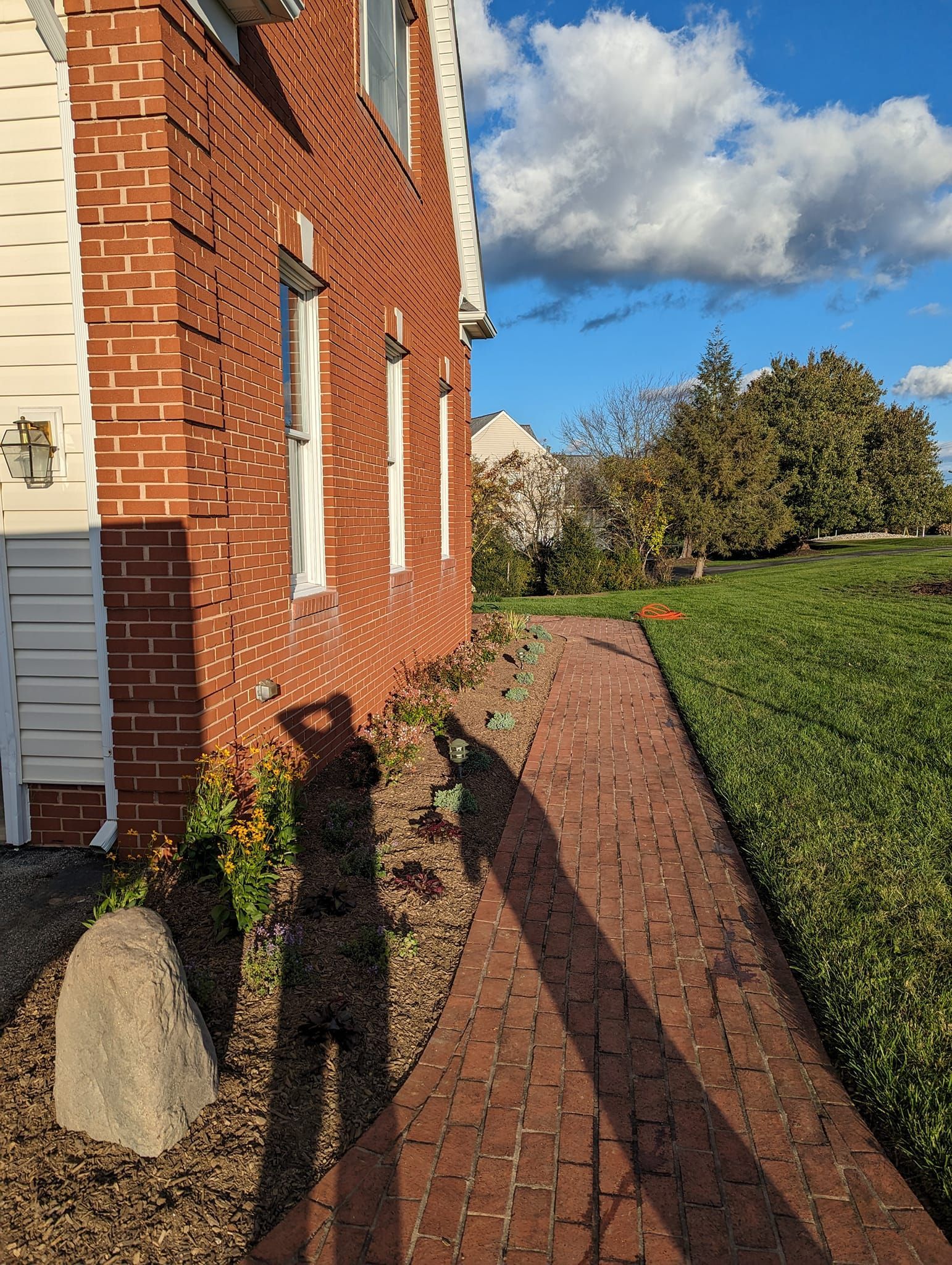 A red brick walkway runs alongside a brick building, with long, human shadows cast across the ground under a sunny sky.