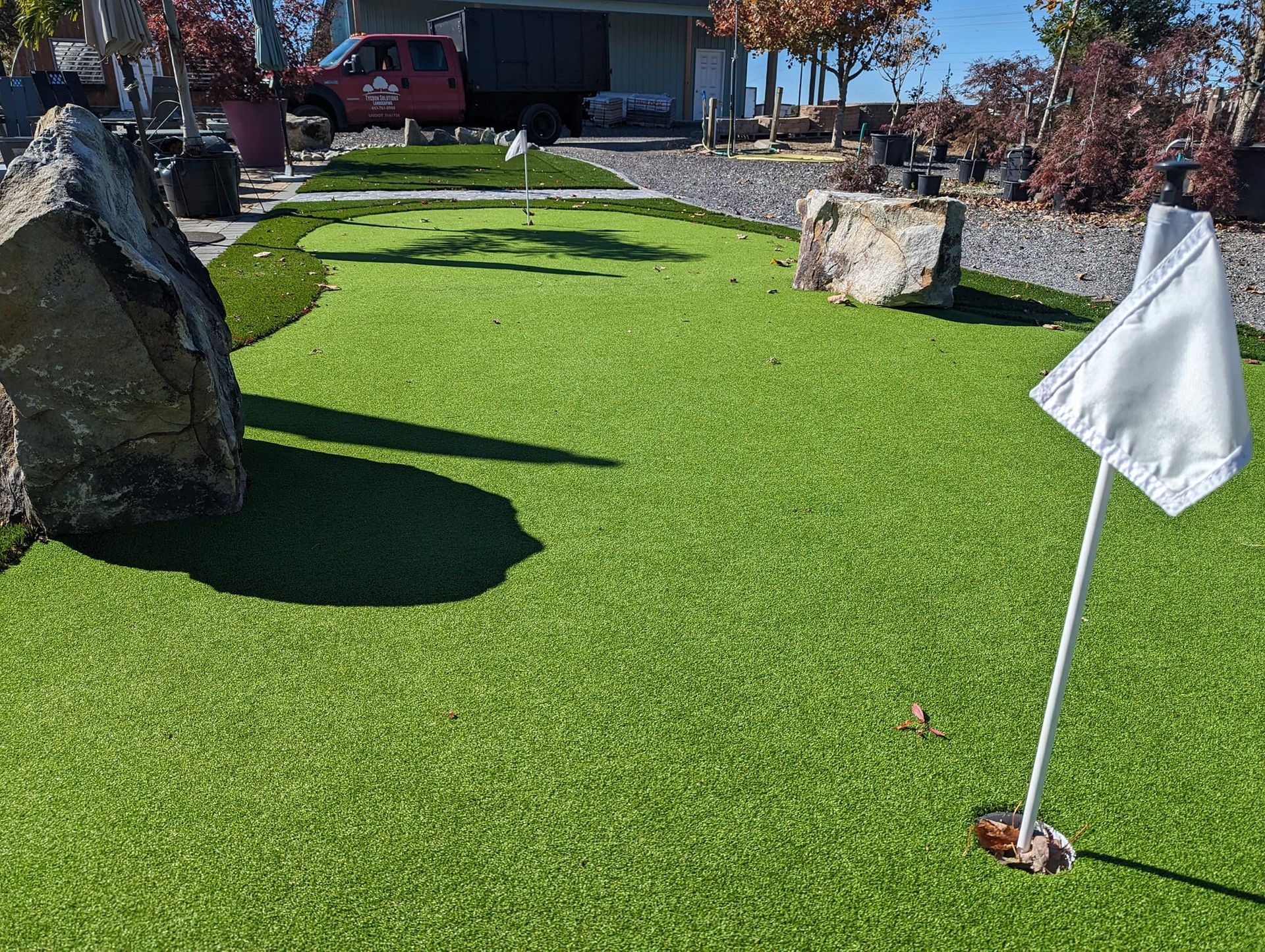 A putting green with a white flag, surrounded by decorative rocks, artificial turf, and a red truck in the background.