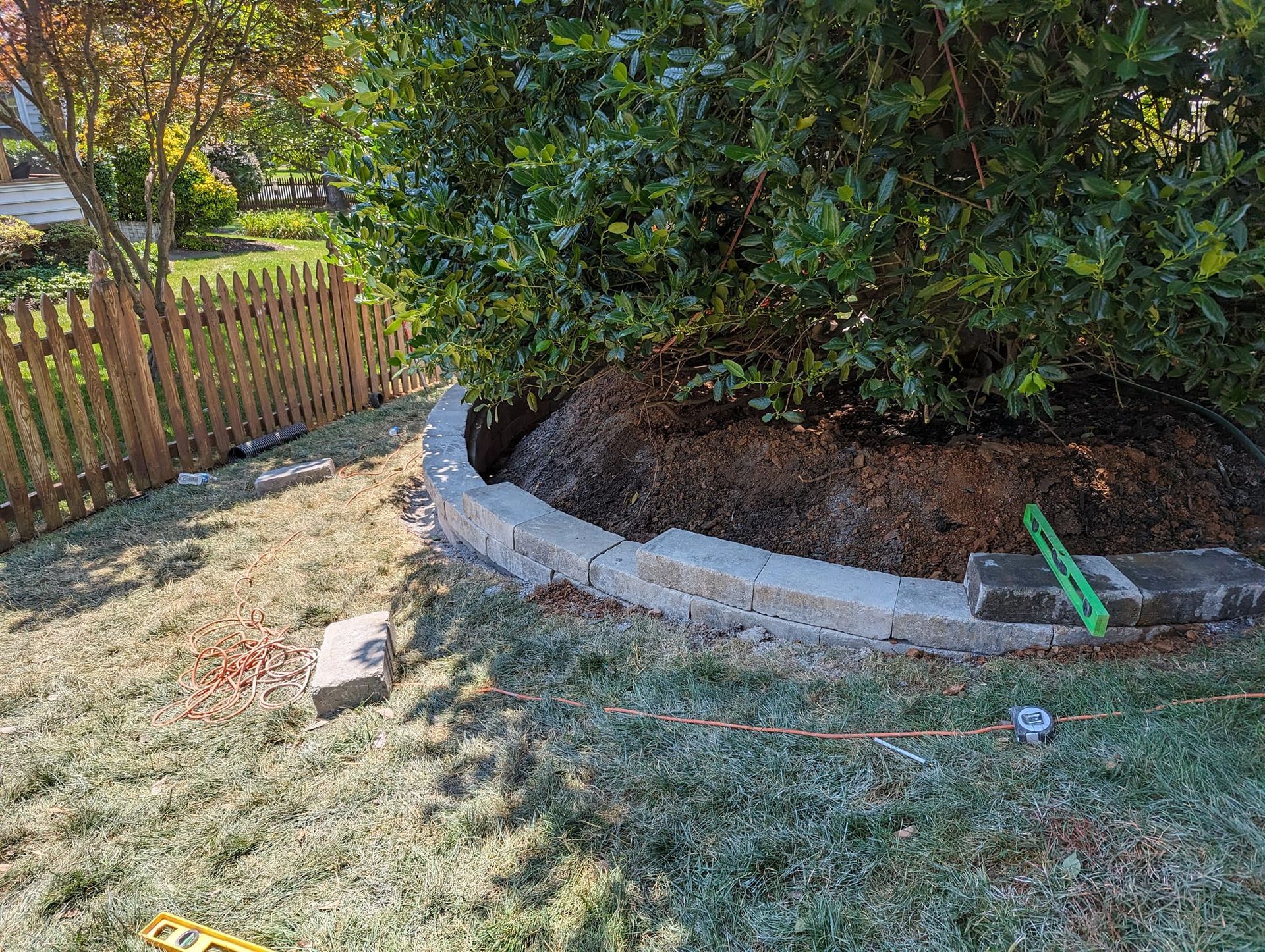 A partially constructed retaining wall made of light-colored stone bricks curves around a large, leafy shrub in a yard.