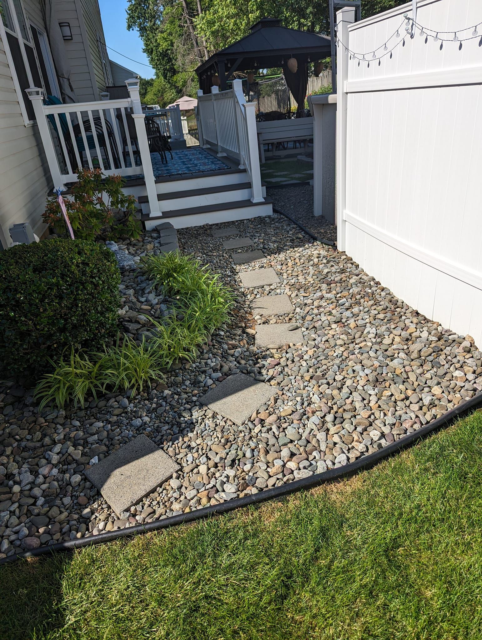 A backyard view showing a white fence, a gravel path with stepping stones, a wooden deck, and a gazebo in the distance.