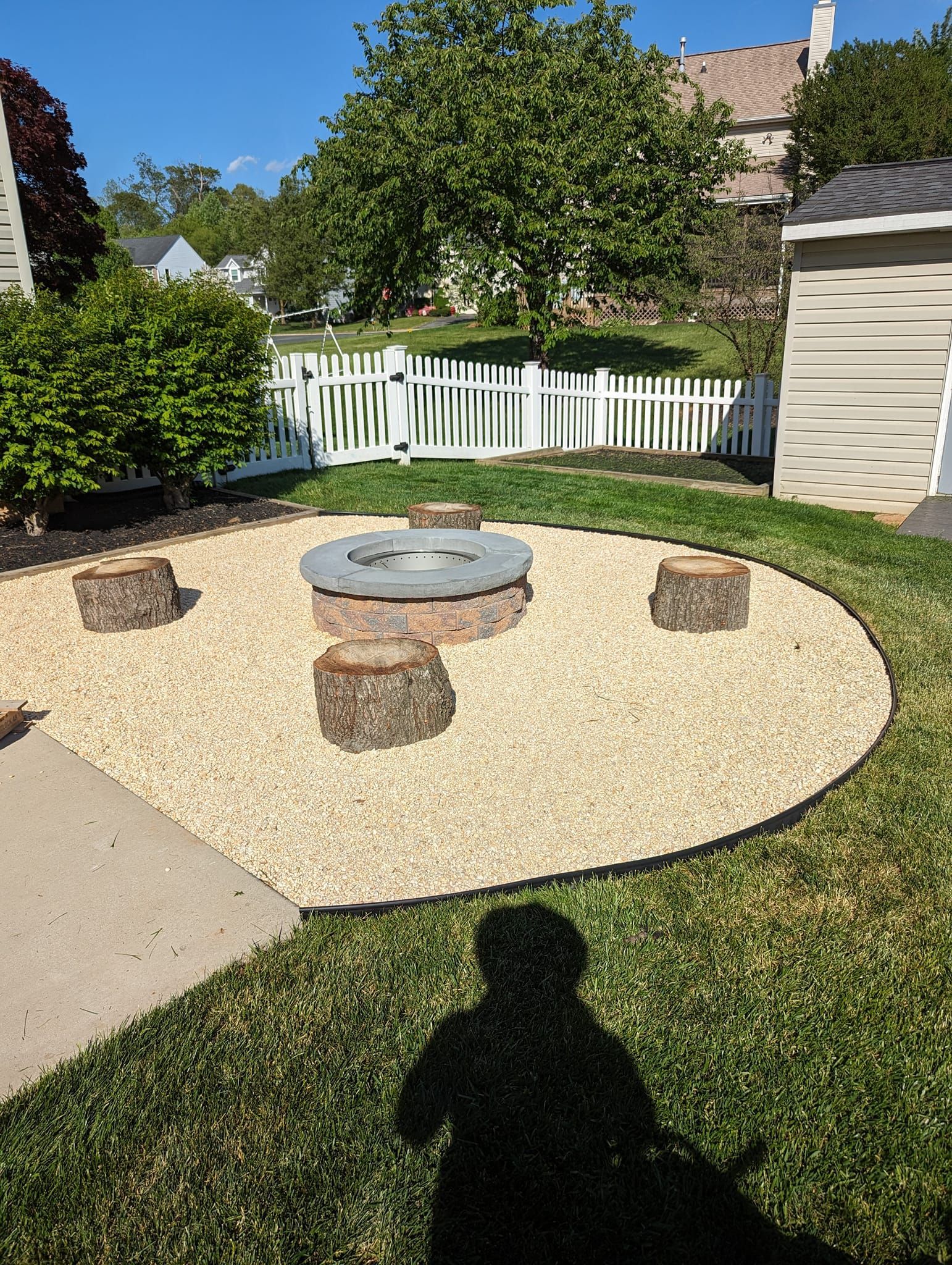 A round gravel fire pit area in a backyard featuring a central stone fire ring surrounded by four wooden log stools.