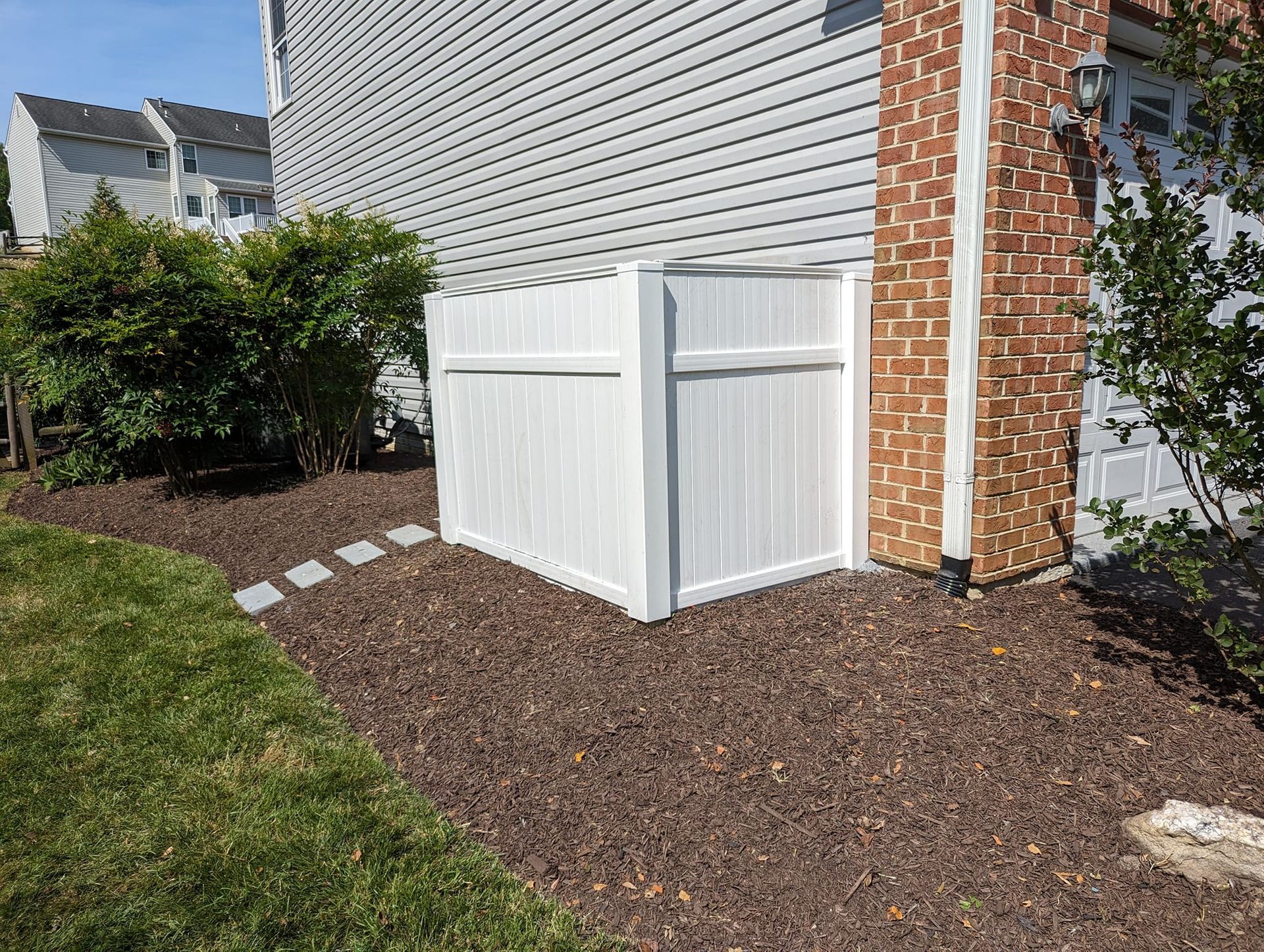 A white vinyl privacy fence screen hides equipment next to a brick-and-siding house, with mulch and a stone path nearby.