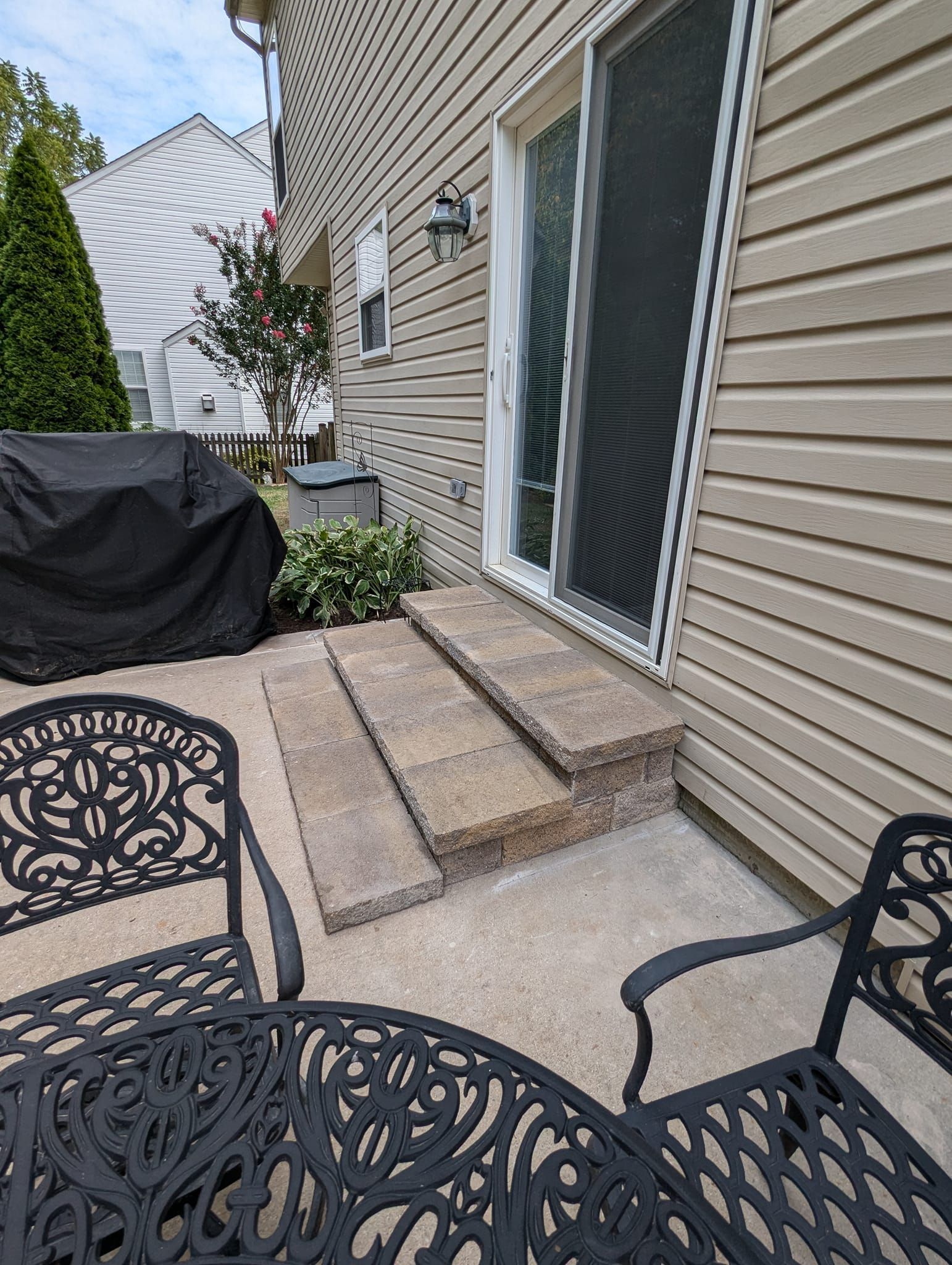A patio with a set of two stone steps leading to a sliding glass door against a tan house, featuring outdoor furniture.