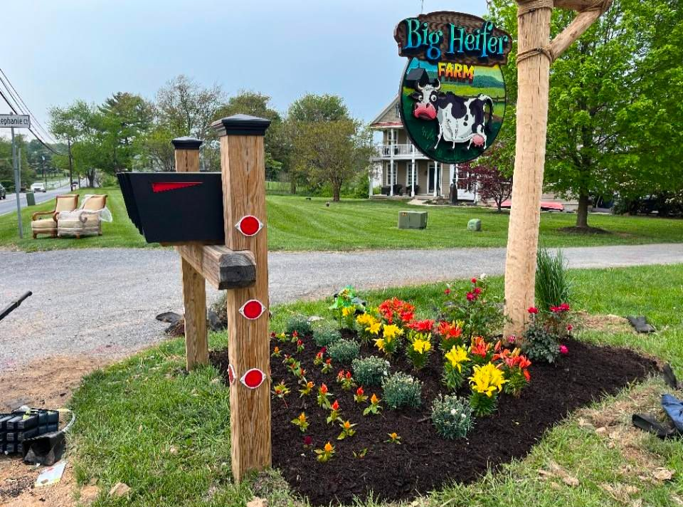 A Big Heifer Farm sign with a cow illustration hangs over a mailbox and a flower bed with mulch in a grassy front yard.