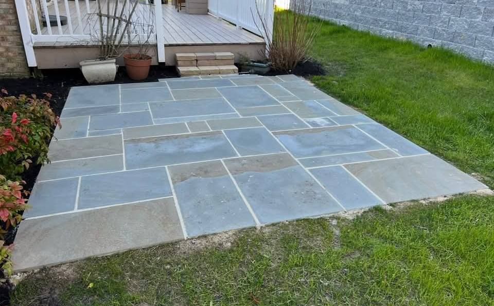 A newly installed gray slate patio made of rectangular tiles, situated in a grassy backyard next to a wooden deck.