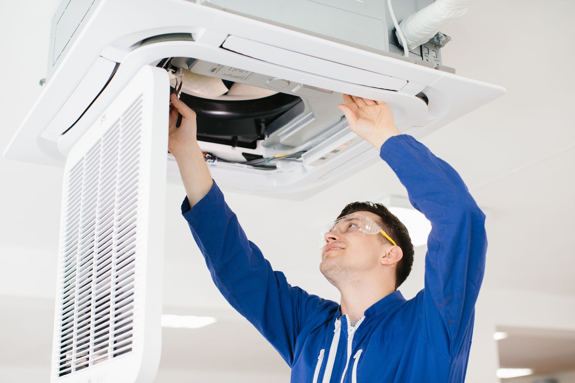 HVAC technician inspecting ceiling-mounted air conditioning unit. Wearing safety glasses and a blue uniform.
