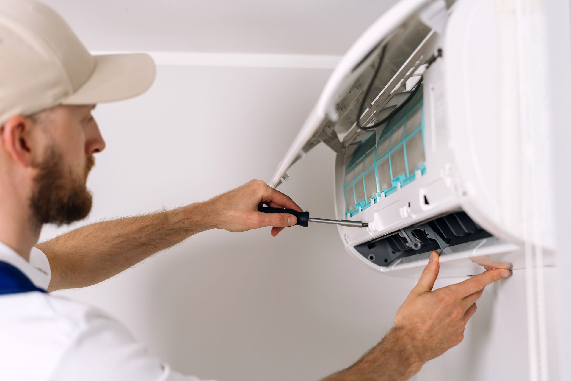Man in cap repairs a wall-mounted AC unit with a screwdriver indoors.