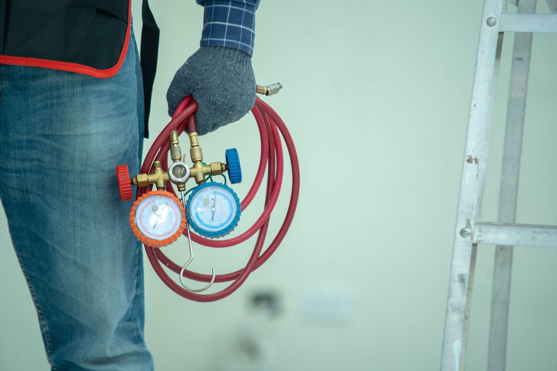 HVAC technician holding gauges and hoses; next to a ladder.
