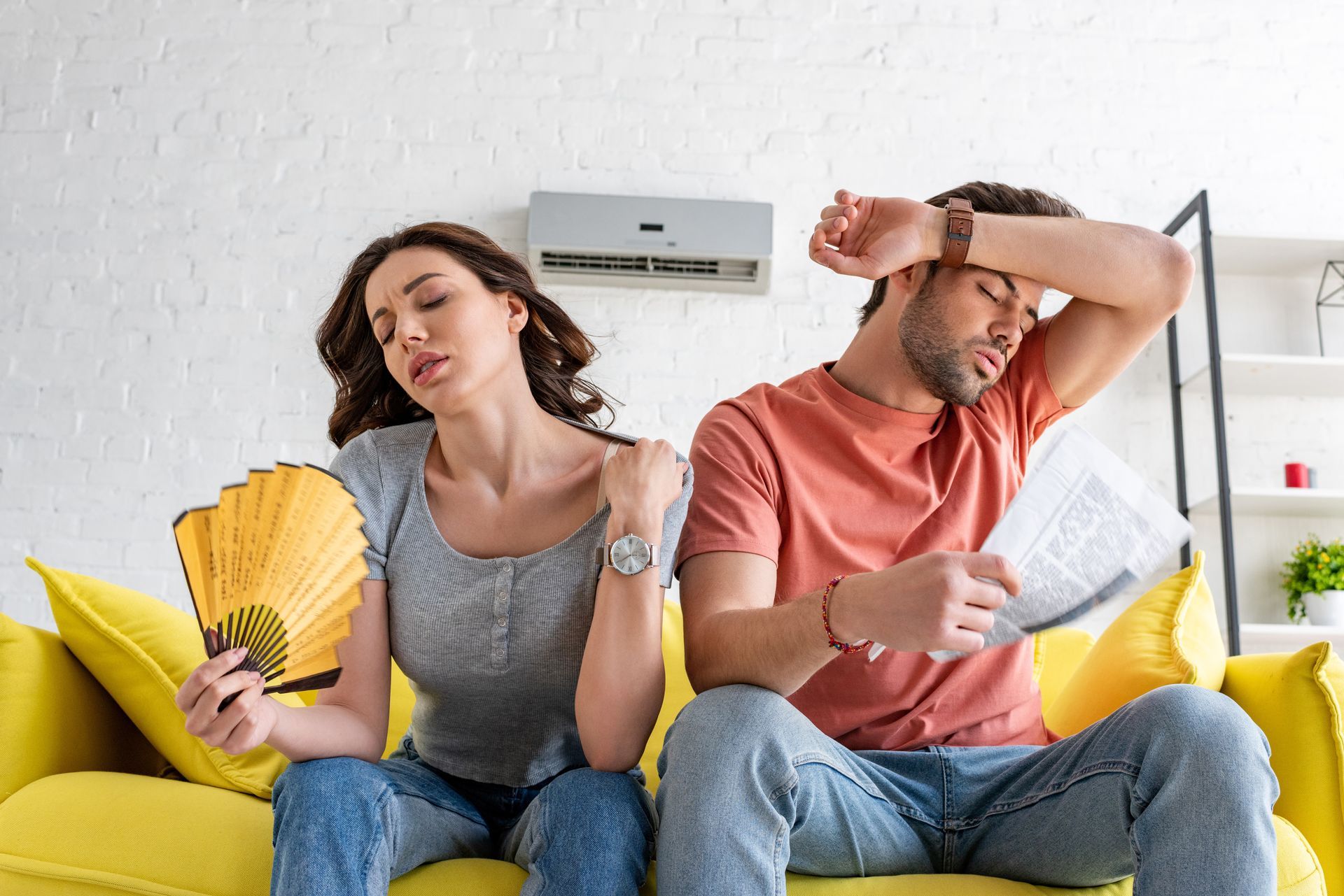 A couple sits on a yellow couch, sweating. Woman uses a fan, man wipes his forehead. Air conditioner on the wall. A couple sits on a yellow couch, sweating. Woman uses a fan, man wipes his forehead. Air conditioner on the wall.