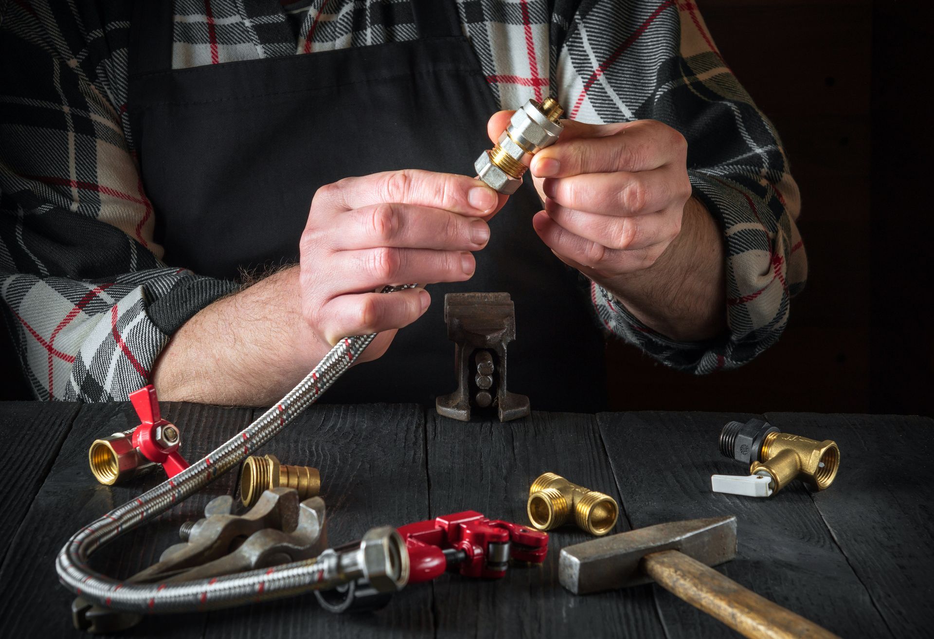 Plumber assembling plumbing parts on a dark wooden table, wearing a plaid shirt and black apron.