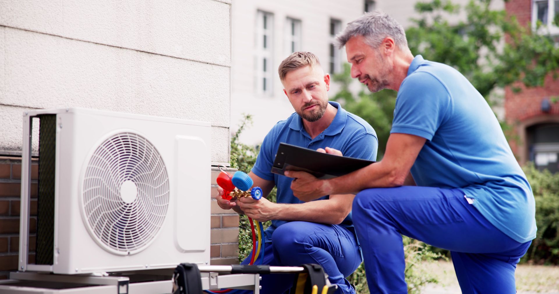 Two technicians in blue uniforms inspecting an outdoor air conditioning unit. One holds a multimeter.