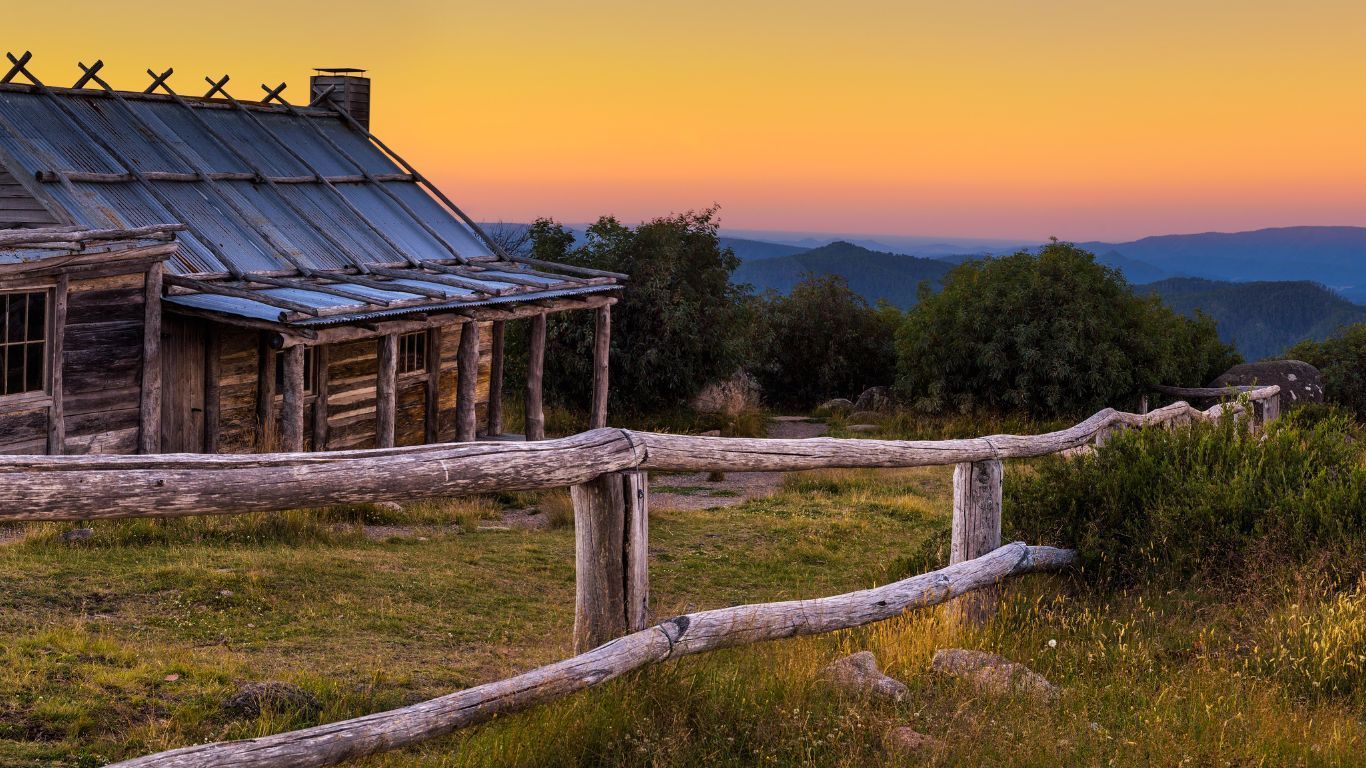 Weathered cabin with a wooden fence overlooks distant hills at sunset.