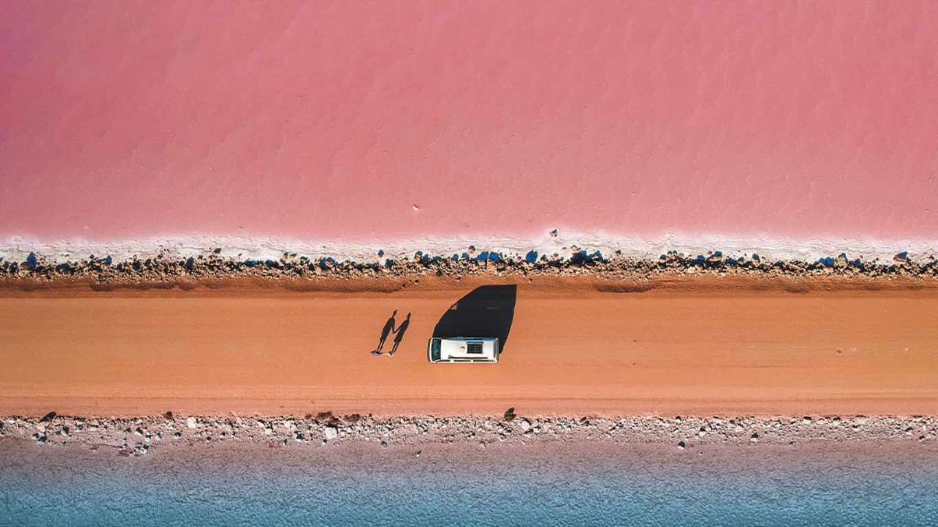 Aerial view: people walking on a red road beside a pink lake and blue ocean.