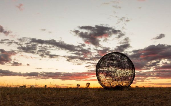 Black, orb-like sculpture in a field at sunset, reflecting the sky.