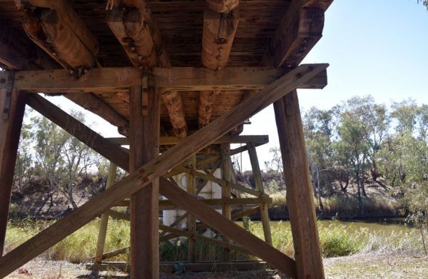 Underside of a weathered wooden bridge over a river, showcasing support beams and cross bracing on a sunny day.