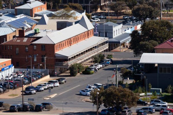 Aerial view of a town street lined with cars and buildings, including a large brick building with a long balcony.