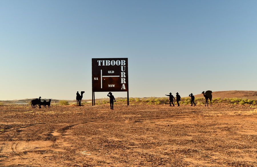 Sign for Tibooburra, Australia, with metal camel sculptures and people in a desert landscape under a clear blue sky.
