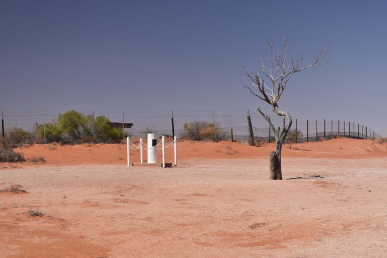 Dry, reddish desert landscape with a leafless tree, a white weather station, and a fenced area under a clear blue sky.