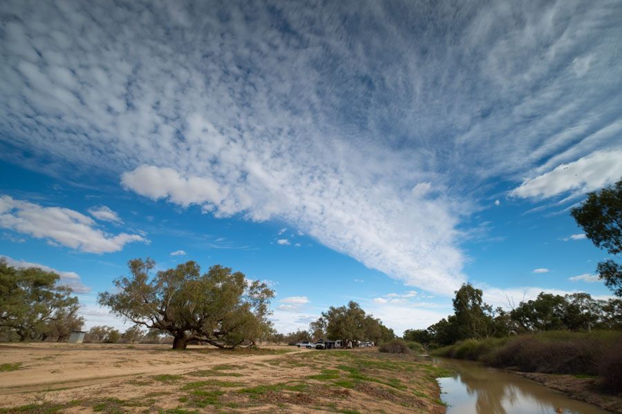 Blue sky with wispy clouds, trees, and a waterway in a dry, open landscape.