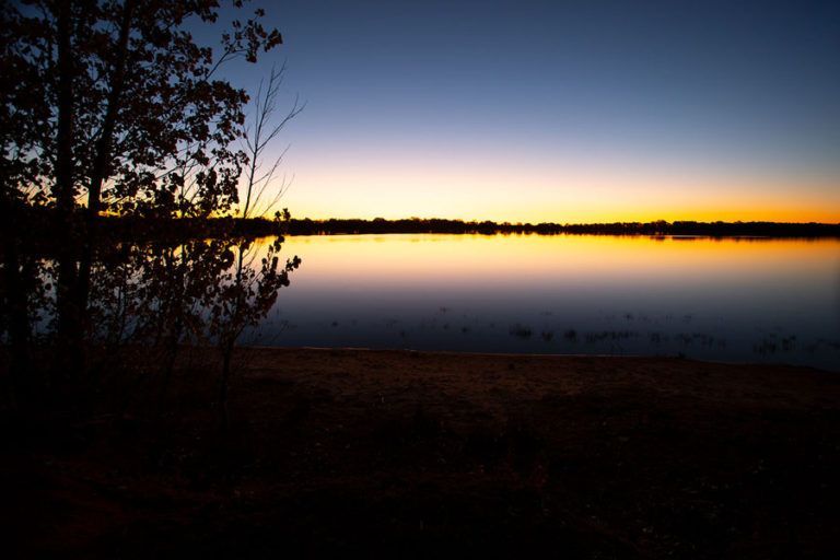 Dusk over a calm lake. Orange and blue sky reflected in the water. Dark silhouettes of trees on the shore.