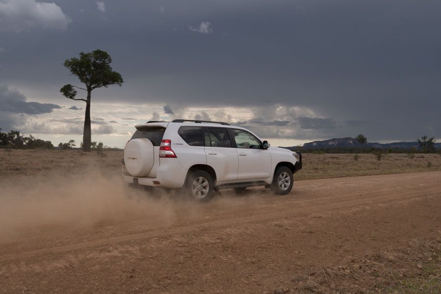 White SUV kicks up dust on a dirt road, under a cloudy sky.