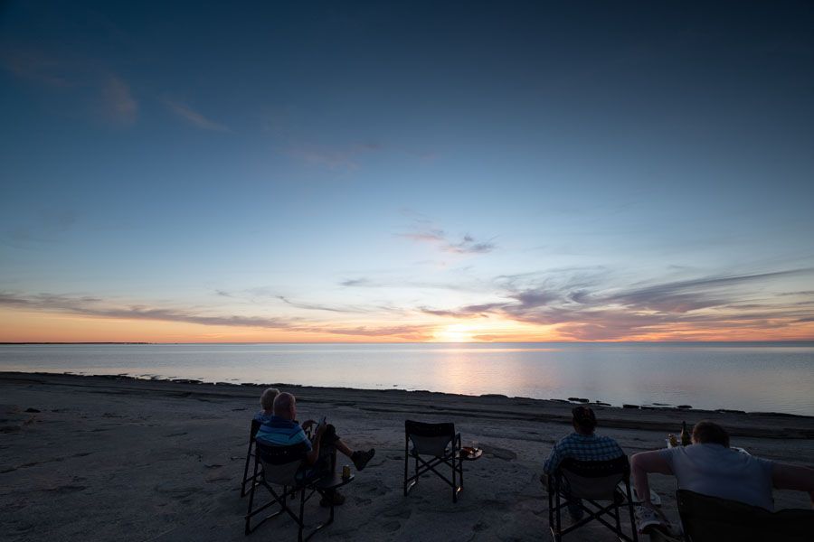 People seated in folding chairs on a beach watching a sunset over the ocean.