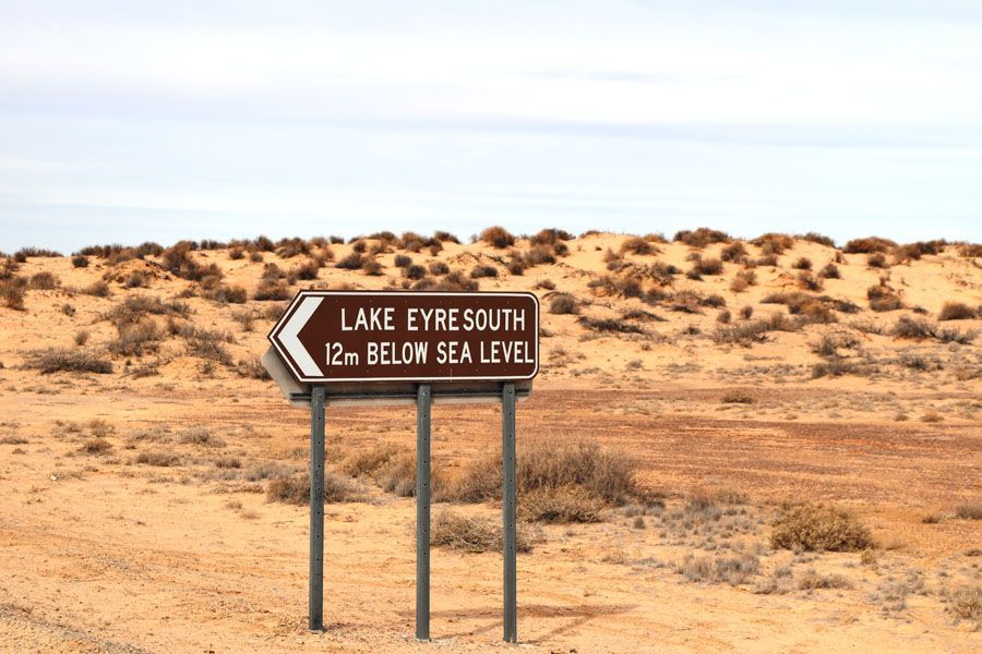 Signpost: Lake Eyre South, 12m below sea level, desert landscape, brown and tan hues.