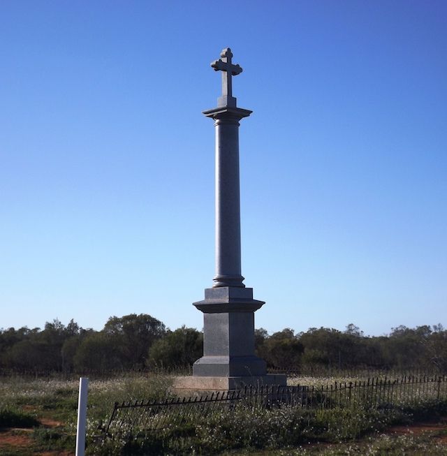 A tall, dark monument with a cross on top, surrounded by a fence and sparse vegetation, under a clear blue sky.