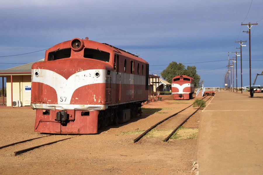 Two red and white trains sit on tracks at a station in a desert landscape.