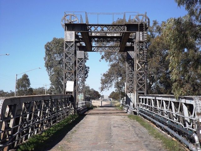 Bridge with metal structure over a dirt road, trees on either side, sunny day.