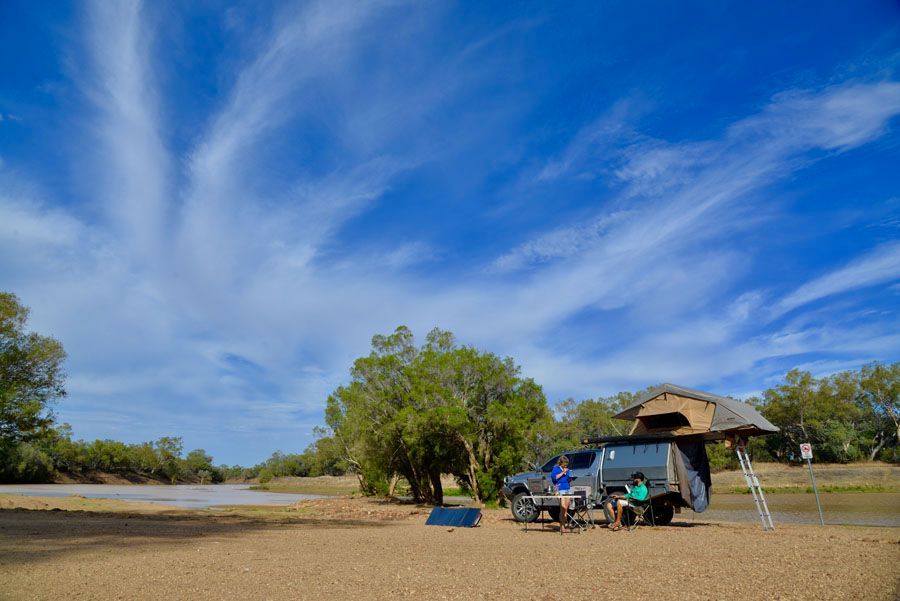 Overlanding campsite by a river, blue sky with wispy clouds. A truck with a rooftop tent and people seated nearby.