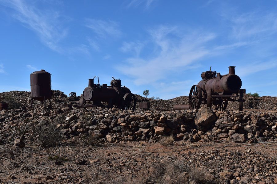 Rusty old steam engines and a tank sit on a rocky plain under a blue sky.