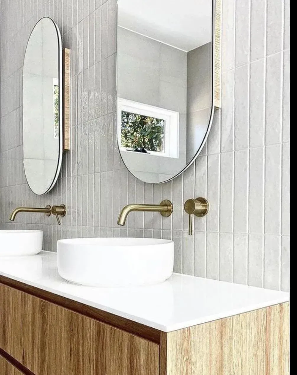 Bathroom with two oval mirrors, gold faucets, white sinks, wooden vanity, and gray vertical tile.