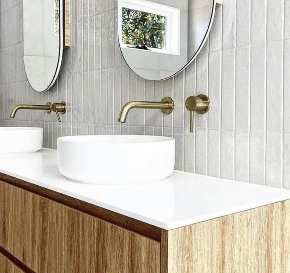 Modern bathroom with dual sinks, gold fixtures, oval mirrors, and light gray tiled walls.