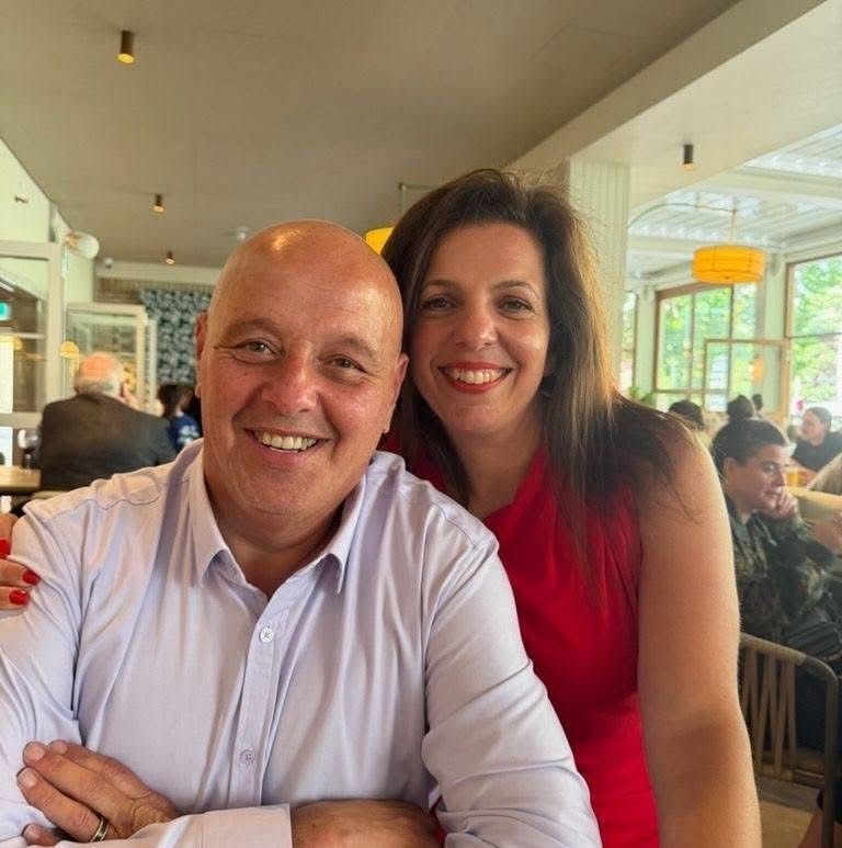 Man and woman smiling, indoors. Man with shaved head in light purple shirt; woman in red dress.