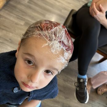 Boy with hair dye under plastic wrap, in a salon, looking up with a concerned expression.