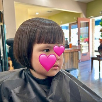 A young child with a new, bowl-shaped haircut and two pink heart emojis covering their face, in a hair salon.