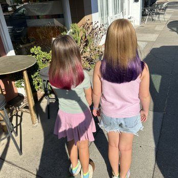 Two young girls, back view, with color-dipped hair, holding hands, outdoors.